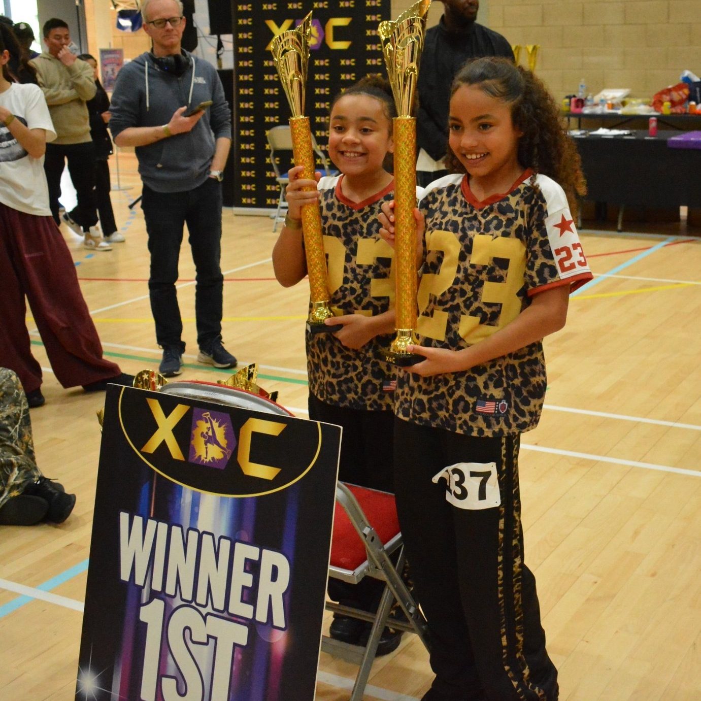 Two Warriorz dancers holding trophies beside an XDC winner board