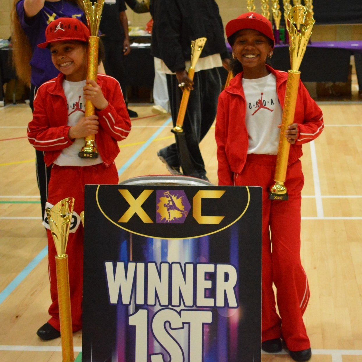 Two Warriorz dancers in red tracksuits with trophies beside an XDC winner board